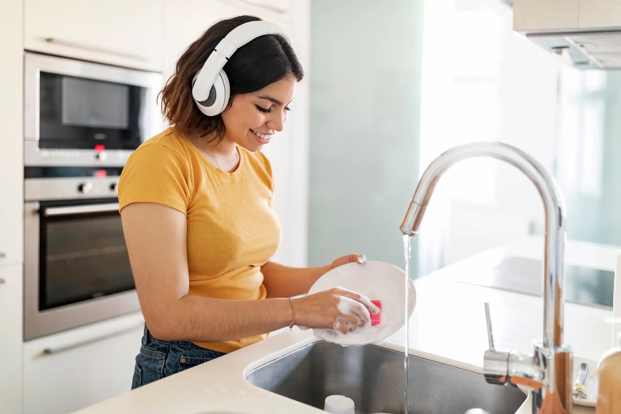 Young Arab Woman Wearing Wireless Headphones Washing Dishes In Kitchen